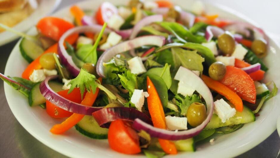 A plate of mixed salad with greens, sliced red onions, bell peppers, olives, cucumber, tomato, and feta cheese.
