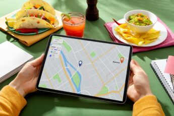 Person holding a tablet displaying a map with a route, surrounded by tacos, chips with guacamole, a drink, and a notebook on a green table.